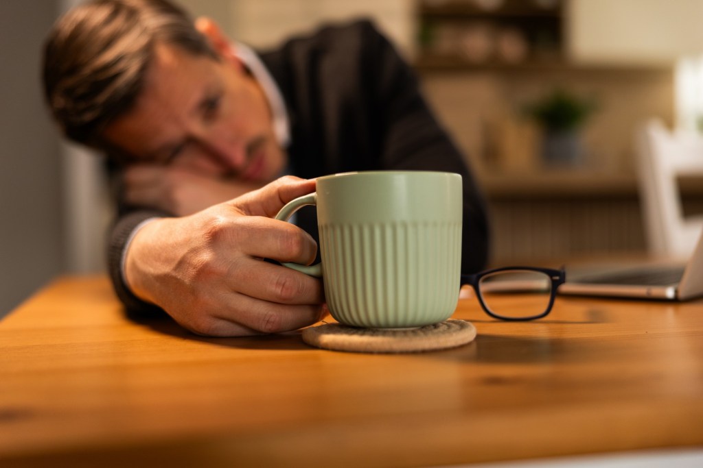 bărbat obosit cu capul pe birou care bea cafea